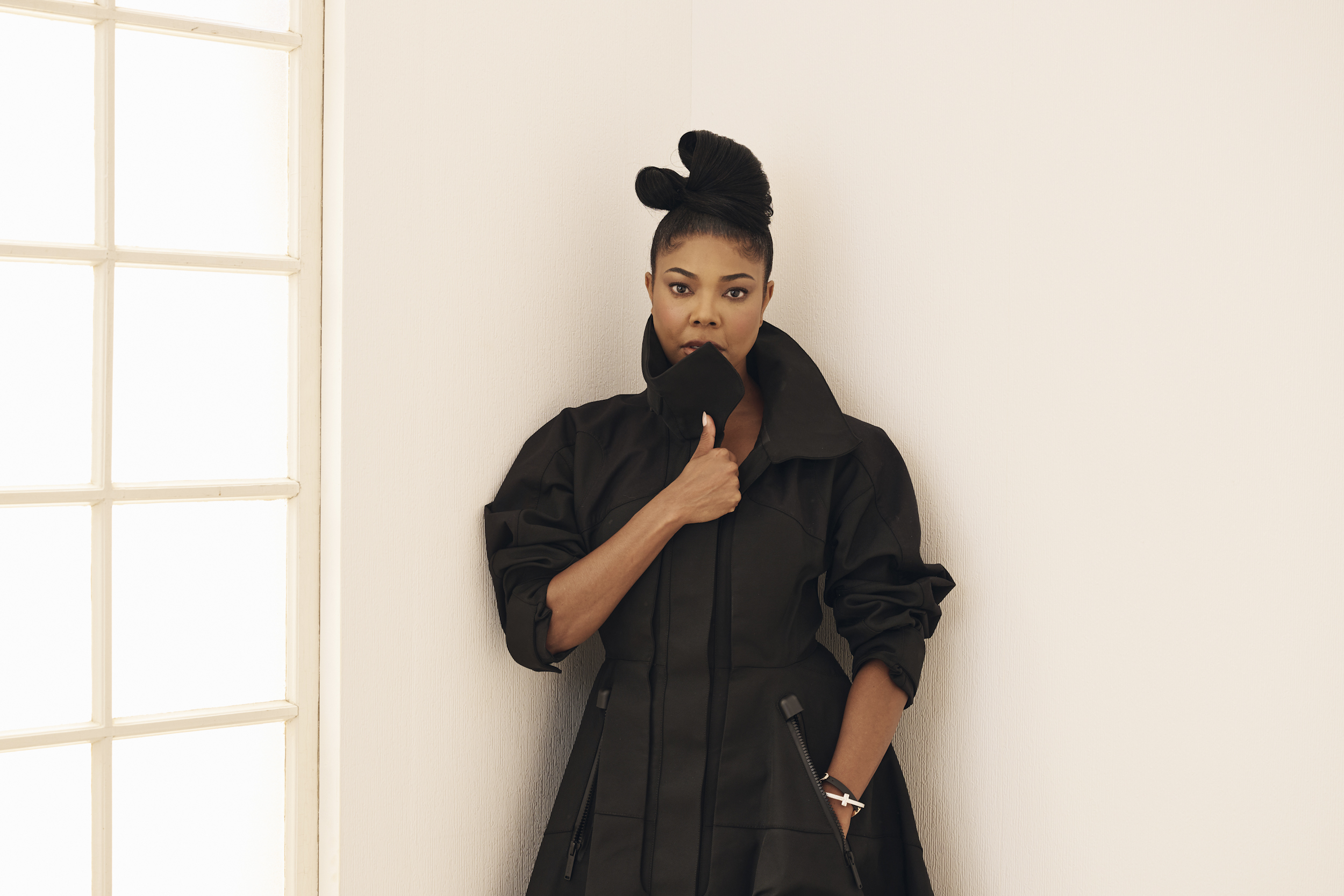 Woman in a black fashion-forward outfit posing against a minimalist white wall, showcasing contemporary design and style, relevant to lifestyle and cultural discussions in 3 Magazine.