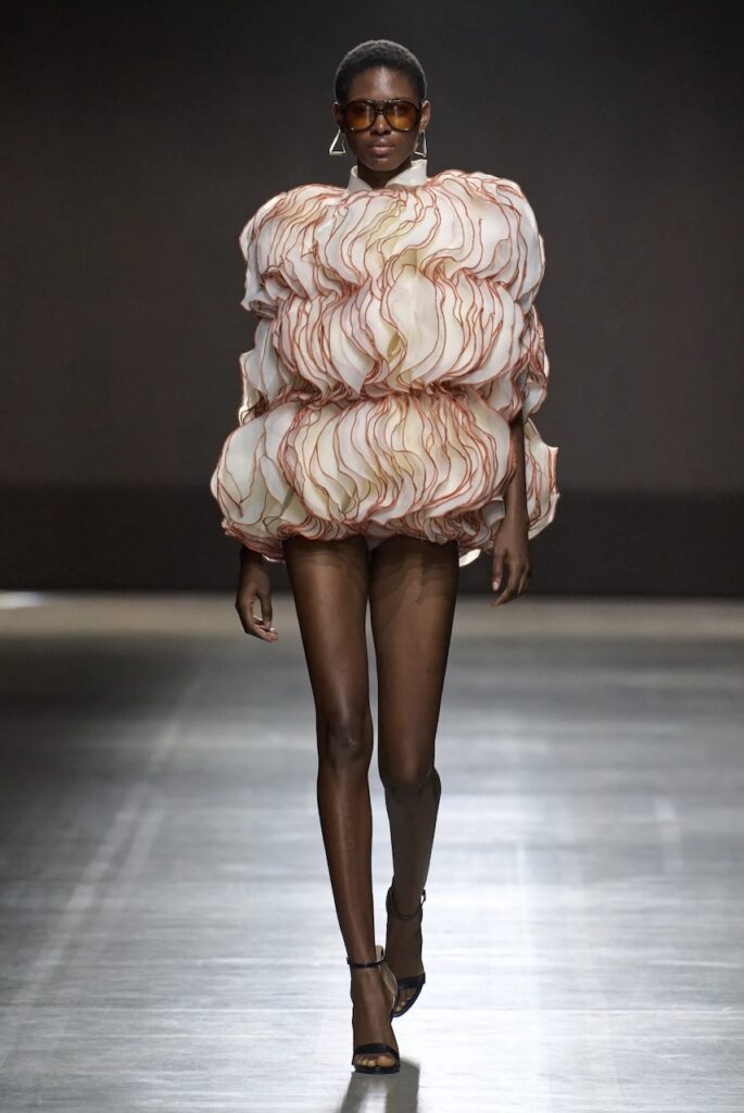 Female model walking down the runway in a ruffled cream mini dress. 