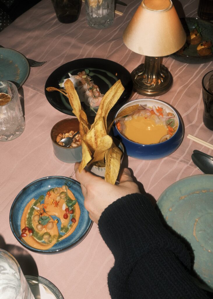 A person holds a cup of plantain chips near a cozy dining table with small plates of colourful dishes.