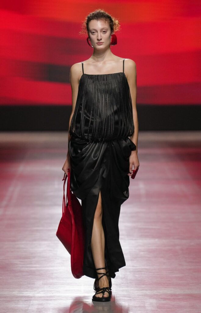 Female model walking down the runway wearing a delicate black dress and holding a large red bag.