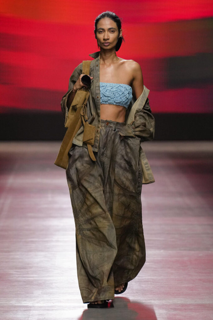 Female model walking down the runway in a light blue tube top with an army green jacket and matching pants.
