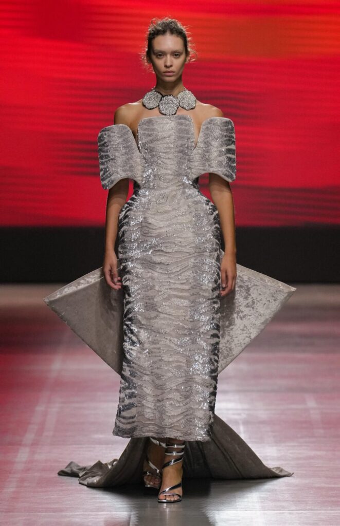 Female model walks down the runway wearing a sculptural grey velvet dress with shiny wavy lines.