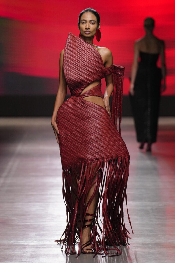 Female model walks down the runway wearing a sculptural red woven dress.
