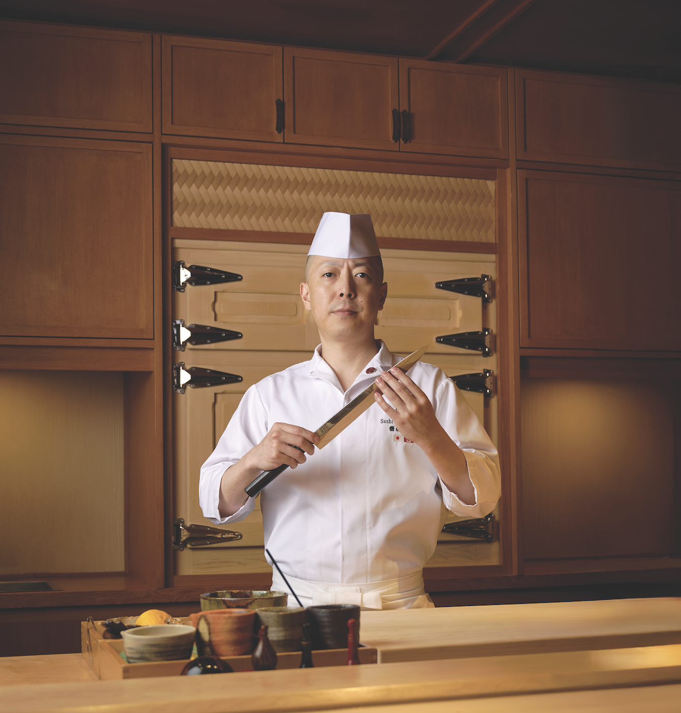 Chef Masaki Saito holding a sushi knife in a traditional Japanese kitchen, surrounded by culinary tools and ingredients, reflecting fine dining and sushi craftsmanship.
