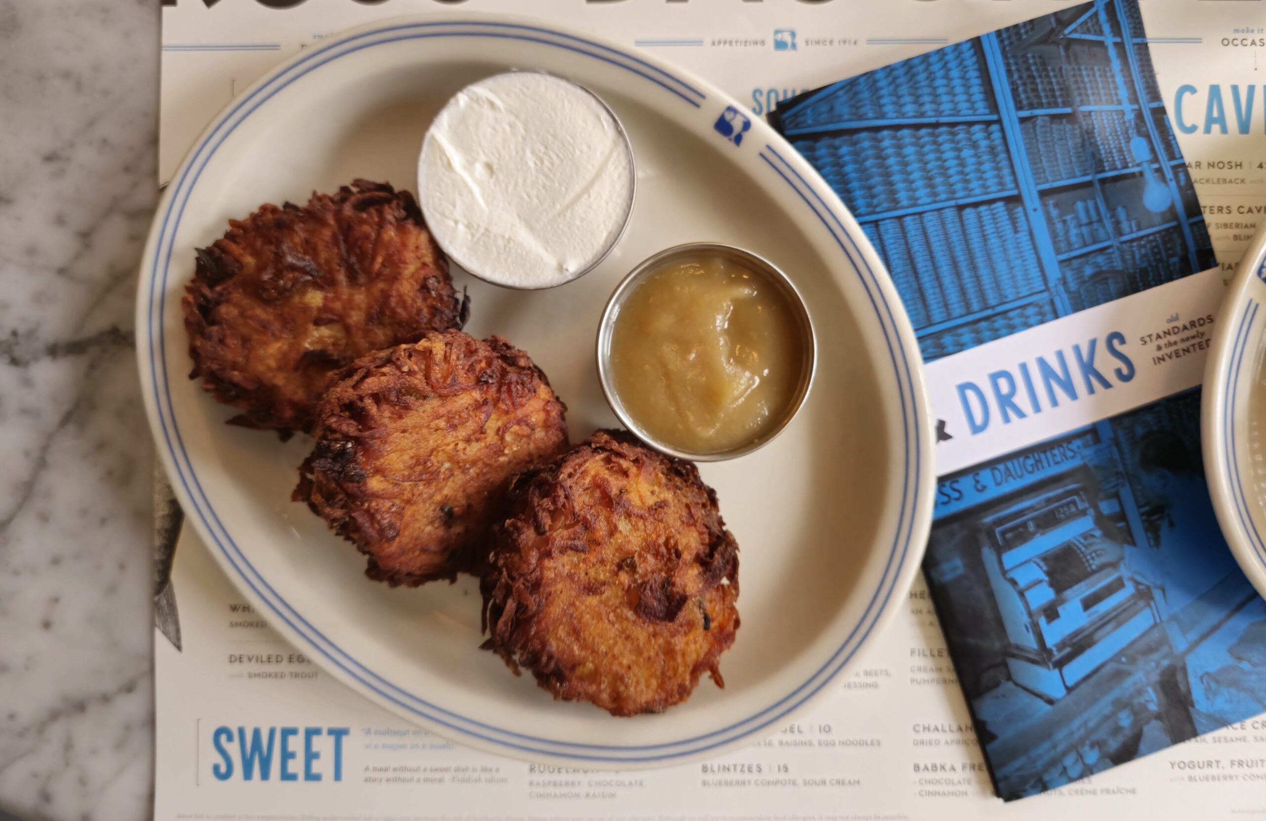Three potato latkes on a plate with two sauces in metal containers. The food and drink menus lay underneath.